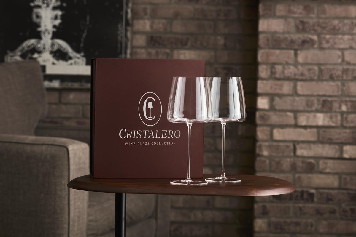 Cristalero wine glass collection with two glasses on a table against a brick wall.