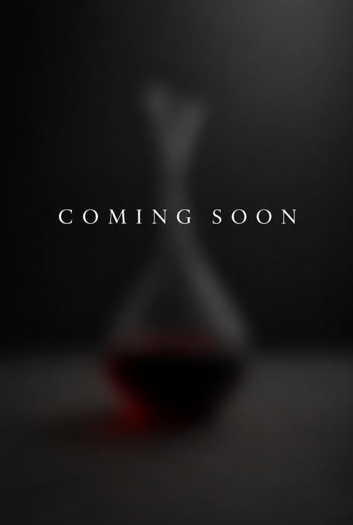 A dark promotional placeholder with the words 'COMING SOON' centered over a blurred glass decanter containing a red liquid on a dark background.